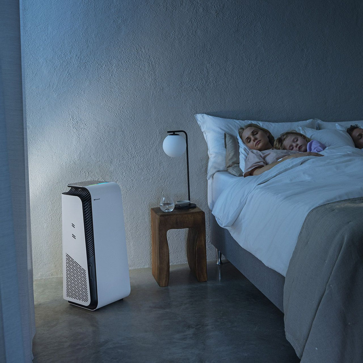 Blueair HealthProtect 7440i Luftreiniger in weiß neben Bett mit schlafender Familie im abgedunkelten Schlafzimmer