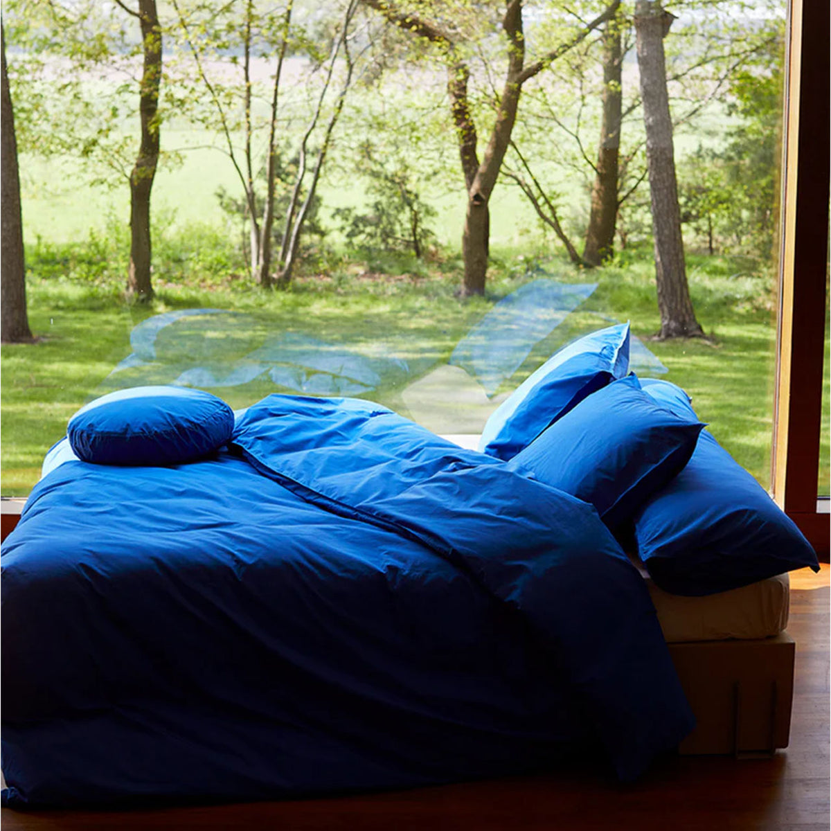 Bett mit Suite702 Perkal Bettdeckenbezug in Cobalt Blue vor großer Fensterfront mit Blick auf Bäume