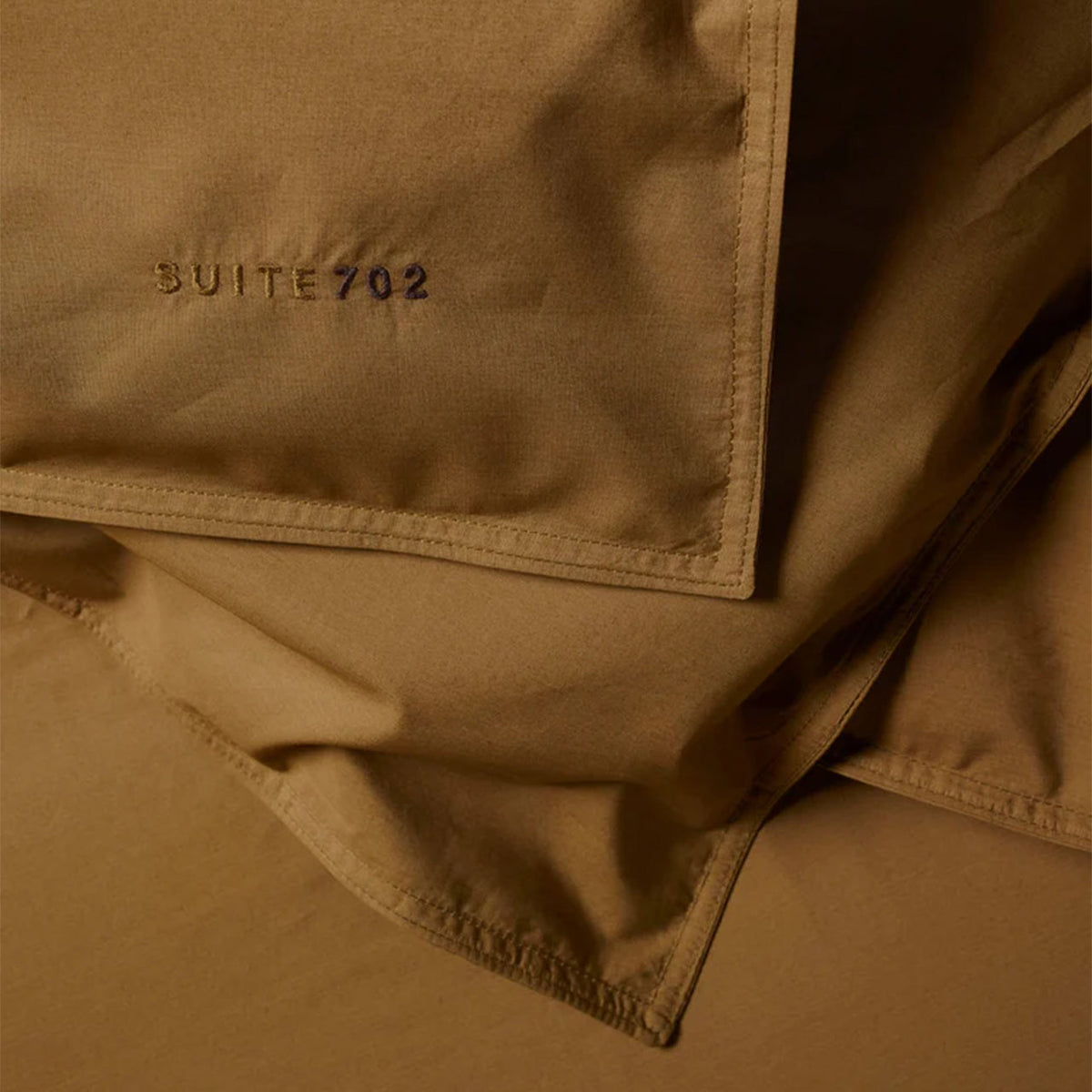 Nahaufnahme der Ecke des Suite702 Perkal Bettdeckenbezugs in Khaki Gold mit gesticktem Schriftzug „Suite702“