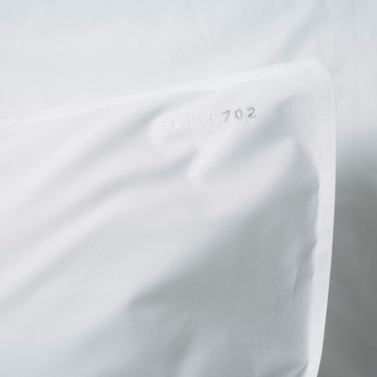 Nahaufnahme der Eckkante des weißen Suite702 Perkal-Kissenbezugs mit eingewebtem Schriftzug „Suite702“